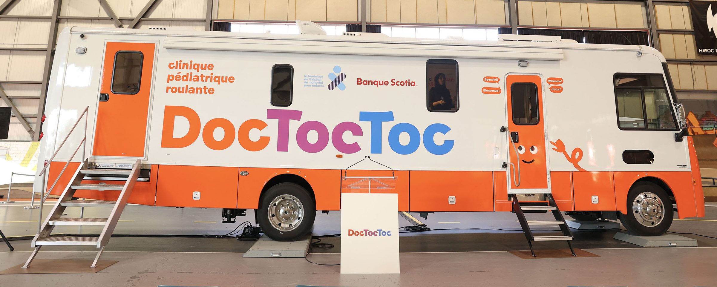 La clinique mobile DocTocToc, autobus orange et blanc, dévoilée sous la charpente du hangar de l'inauguration, le 19 mars 2025